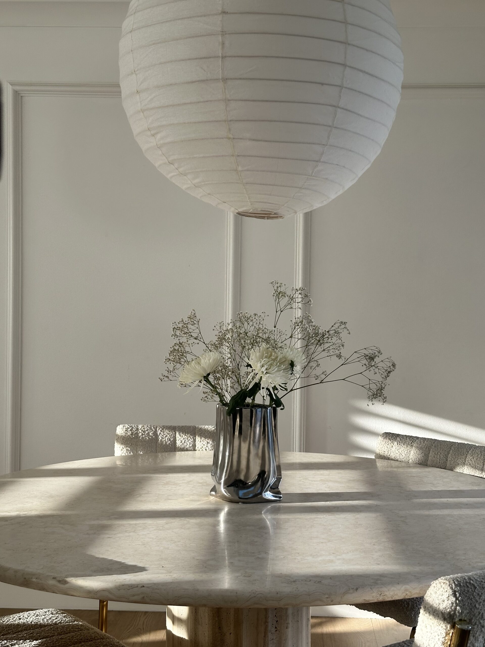 A serene, light-filled home dining area, featuring a round marble table, textured chairs, and a minimalist paper pendant light, showcases a vase of white flowers. This elegantly designed space demonstrates how a personal environment becomes your ultimate business asset for creative entrepreneurs, fostering inspiration and serving as a living portfolio for brand alignment and business success.