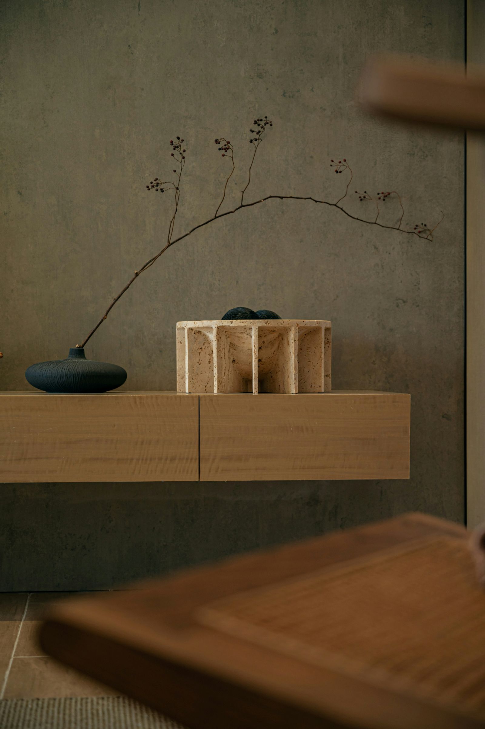 A serene and sophisticated unhurried interior vignette, showcasing a floating oak cabinet set against a textured plaster wall. The arrangement features a dark, organic vase with a delicate branch and a sculptural stone object, highlighting natural materials, subtle textures, and the enduring beauty of deliberate design.
