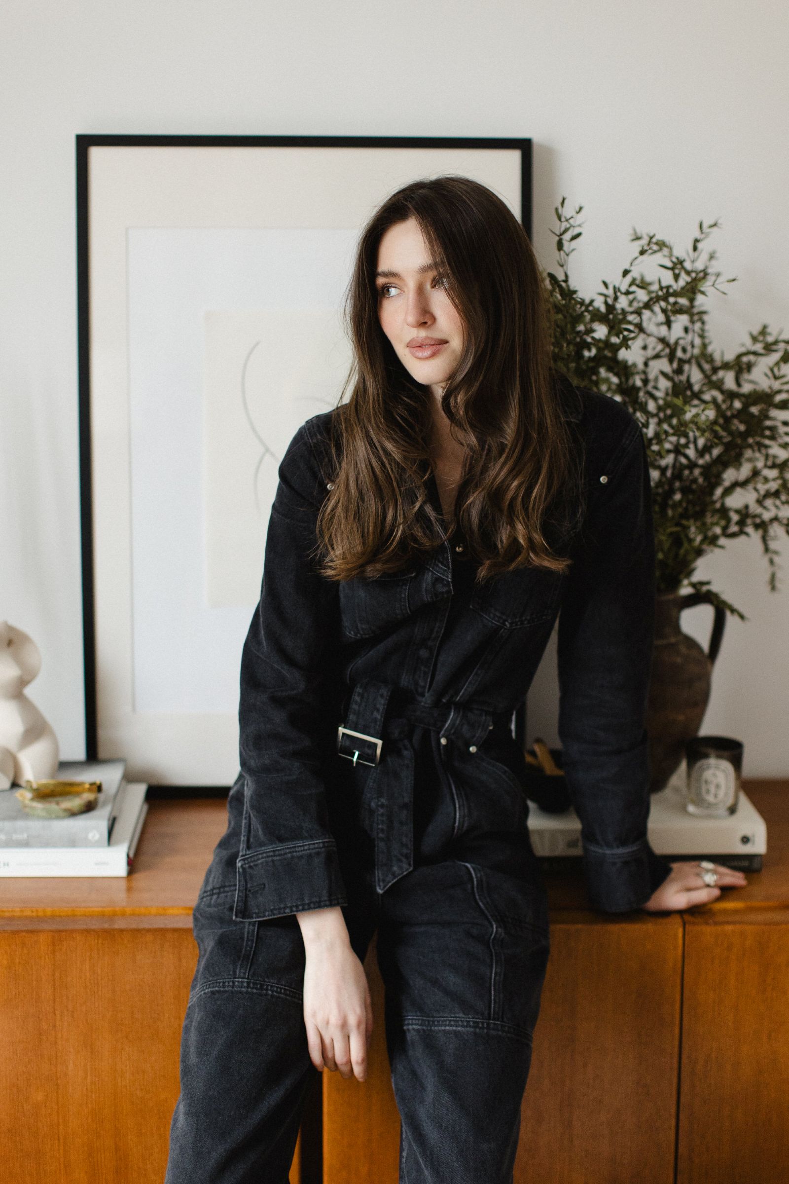 A woman in a dark indigo denim jumpsuit leans against a midcentury wooden sideboard, her posture relaxed, gaze turned away from the lens. Behind her, a composed vignette unfolds—framed artwork, a clay vessel filled with fresh greenery, stacked books, tapered candles, palo santo, and collected objects—styled with an effortless sense of balance and intent.