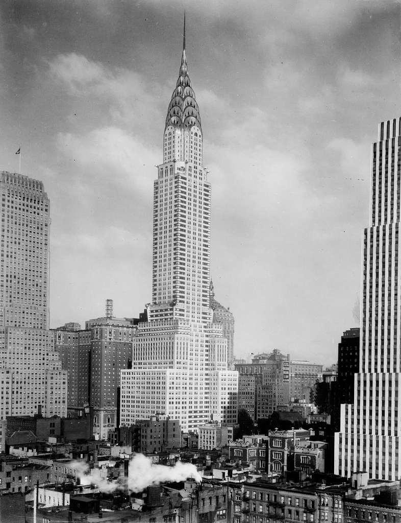 A historic black and white photograph showcasing the majestic Art Deco Chrysler Building soaring above the New York City skyline. Its distinctive geometric spire and streamlined form exemplify the innovative architecture, glamour, and machine age aesthetic of the 1920s design movement.