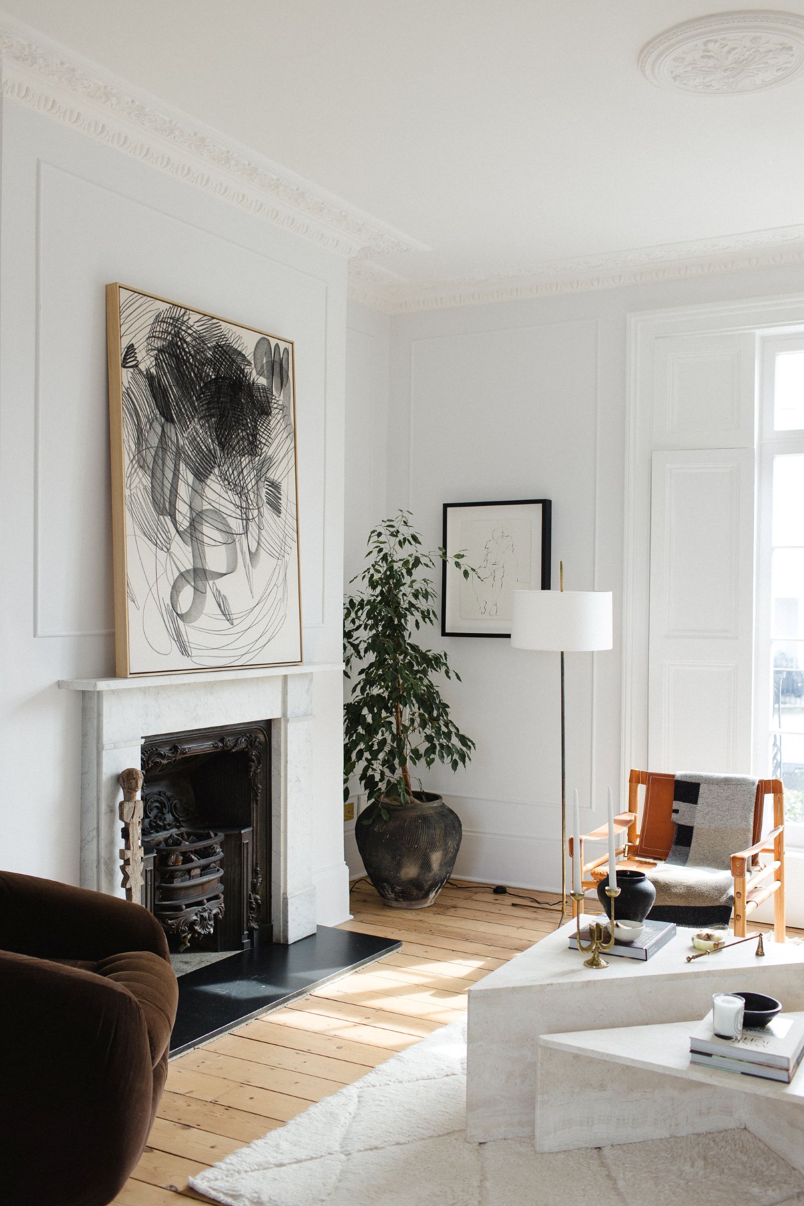 A contemporary, light-filled living room featuring a minimalist white fireplace with a striking abstract artwork above, demonstrating the thoughtful application of design education in luxury residential interiors. The space is softened by a large indoor ficus plant, a plush brown velvet armchair, a stylish wooden and upholstered lounge chair, and a low white coffee table adorned with subtle decor. The light wooden floorboards and high ceilings contribute to a serene, human-centric environment that exemplifies how professional design education cultivates expertise to craft spaces focused on wellbeing and refined living.
