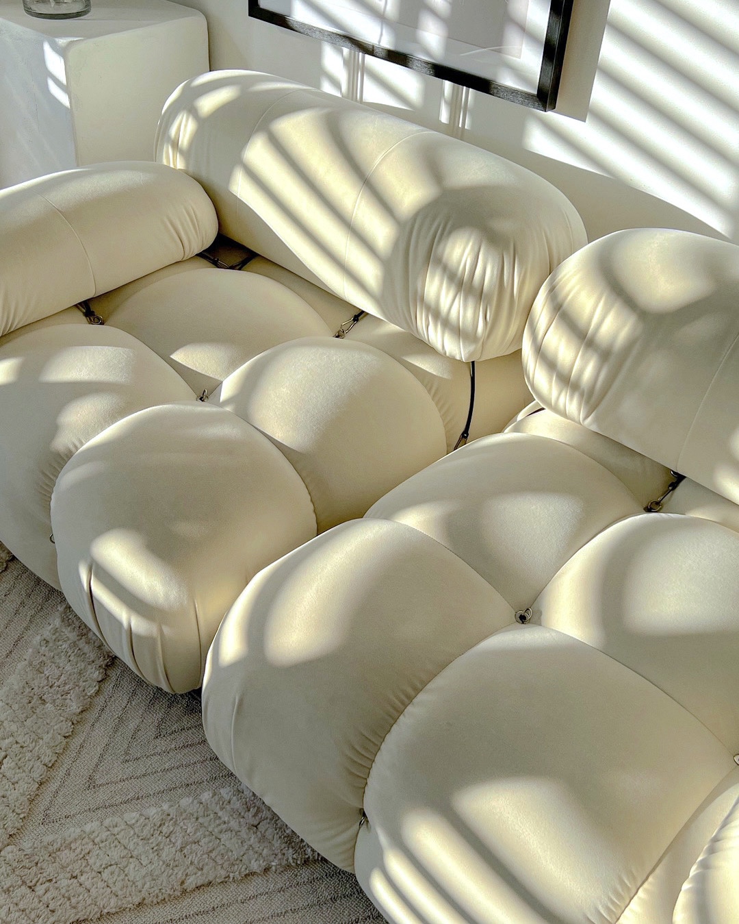 A close-up of a luxurious, cream-coloured modular sofa (reminiscent of a Mario Bellini Camaleonda) bathed in soft, dappled natural sunlight. The interplay of light and shadow highlights the plush, textured upholstery, illustrating how natural light influences the perception of neutral colours and enhances the atmospheric and emotional resonance within an interior space, aligning with principles of colour psychology for wellbeing.