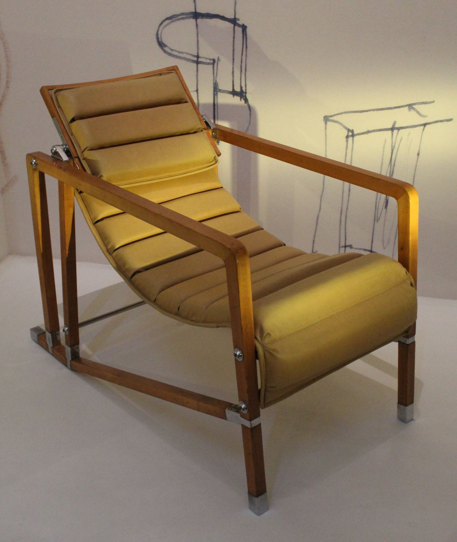A full view of the Transat armchair by Eileen Gray, featuring a horizontally ribbed, gold-toned upholstered sling seat and back, supported by a rich wooden frame with sleek metal brackets, displayed in an exhibition setting with architectural sketches in the background.